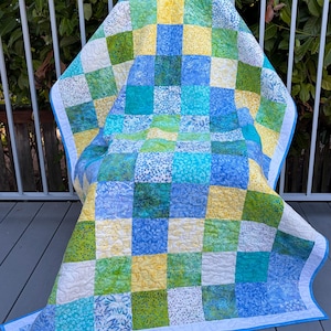 Peut inclure: Une courtepointe en patchwork composée d'une grille de carrés de tissu dans des tons de jaune, vert lime, turquoise et bleu. Le tissu présente divers motifs floraux et botaniques. La courtepointe est finie avec une bordure bleu clair.