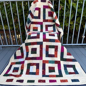 May include: A quilt featuring a repeating block pattern of cream-coloured squares framed by multi-coloured fabric strips in shades of deep red, forest green, navy blue, and magenta. The fabric has a textured appearance with intricate machine quilting.