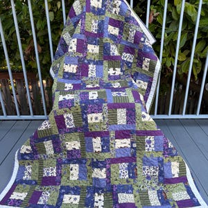 May include: A patchwork quilt featuring a repeating block pattern in shades of purple, olive green, and cream. The fabric squares display various floral, striped, and butterfly prints, finished with a white border and intricate machine quilting.