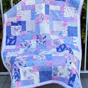 Peut inclure: Un quilt en patchwork composé de divers blocs de tissu carrés dans des tons de bleu, lavande et blanc. La courtepointe présente des motifs floraux, des épis de blé et des imprimés géométriques avec des surpiqûres complexes et une bordure rose clair.