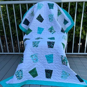 May include: A white quilt with a turquoise border and diamond-shaped patterns in shades of green and turquoise. Some patterns feature floral designs. The quilt is draped over a chair on a wooden deck.