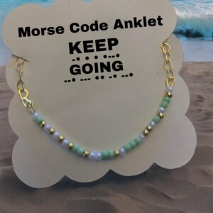 May include: A gold chain anklet with green and white beads. The anklet is on a white card with the text "Morse Code Anklet KEEP GOING..." in black.