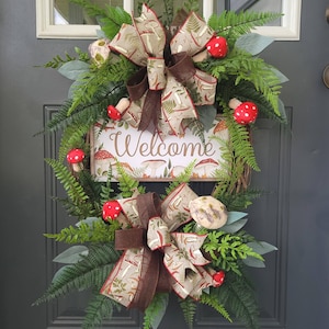 May include: A decorative wreath with a "Welcome" sign, featuring a mushroom theme. The wreath includes green fern-like leaves, red and white spotted mushroom accents, and two large bows with mushroom and fern patterns. The sign has a mushroom border.