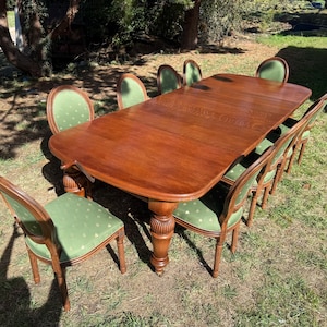 May include: A large rectangular wooden dining table with rounded corners and thick, carved pedestal legs, accompanied by eight matching chairs featuring wooden frames and green upholstered seats with a subtle, repeating geometric pattern.