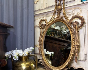 Puede incluir: Un gran espejo ovalado adornado, pintado en oro, con un marco tallado decorativo. El espejo está colgado en una pared y se refleja en el vidrio.