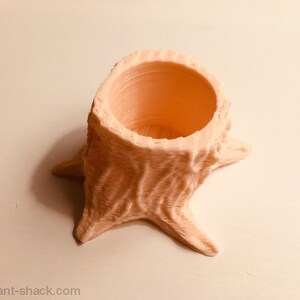 Op de afbeelding: Een kleine, lichtroze, 3D-geprinte plantenpot in de vorm van een boomstronk. De pot heeft een gladde, ronde opening aan de bovenkant.
