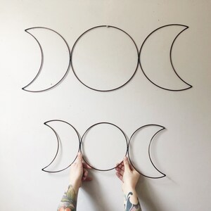 May include: Two metal wall hangings in the shape of the Triple Moon symbol, with a central circle flanked by crescent moons. The top piece is mounted on a light-colored wall. The bottom piece is being held by two hands.