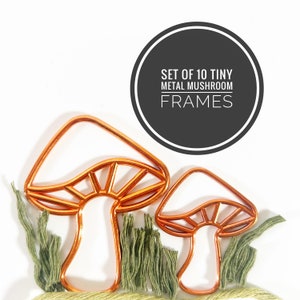 May include: Two copper-colored metal mushroom frames with green embroidered grass accents. The image includes a black circle with the text "SET OF 10 TINY METAL MUSHROOM FRAMES". The mushroom frames are a whimsical decorative item.