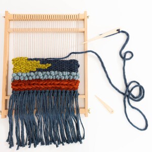 K&ouml;nnte beinhalten: Ein Holzwebstuhl mit einem gewebten Wandteppich mit einem gelben, blauen und orangefarbenen Design. Der Wandteppich hat unten einen Fransen. Ein blaues Garn ist um den Webstuhl gewickelt und zwei h&ouml;lzerne Webnadeln sind enthalten.