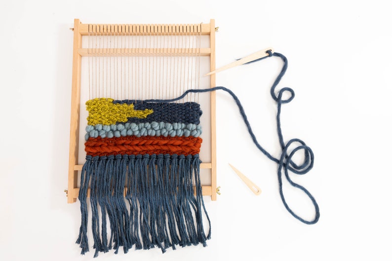 K&ouml;nnte beinhalten: Ein Holzwebstuhl mit einem gewebten Wandteppich mit einem gelben, blauen und orangefarbenen Design. Der Wandteppich hat unten einen Fransen. Ein blaues Garn ist um den Webstuhl gewickelt und zwei h&ouml;lzerne Webnadeln sind enthalten.