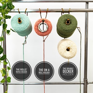 May include: Four spools of yarn are hanging from a metal rack. The spools are green, pink, white, and beige. Each spool has a different type of yarn holder attached to it. The holders are labeled "The Rolls' Royce Holder", "The On A Roll Holder", and "The Double Decker Holder".