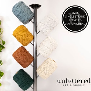 May include: Six spools of 7mm single strand recycled cotton string in various colors, including teal, mustard yellow, burnt orange, white, and forest green, displayed on a metal stand. The label reads "7MM SINGLE STRAND RECYCLED COTTON STRING".