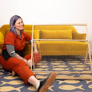 K&ouml;nnte beinhalten: Eine Person sitzt auf einem blauen und gelben Teppich mit Fischmuster und arbeitet an einem h&ouml;lzernen Webstuhl. Der Webstuhl ist vor einem gelben Sofa aufgestellt. Im Hintergrund befinden sich viele Wollkn&auml;uel in verschiedenen Farben.