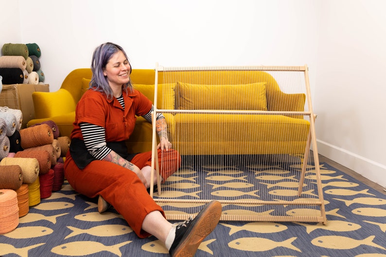 K&ouml;nnte beinhalten: Eine Person sitzt auf einem blauen und gelben Teppich mit Fischmuster und arbeitet an einem h&ouml;lzernen Webstuhl. Der Webstuhl ist vor einem gelben Sofa aufgestellt. Im Hintergrund befinden sich viele Wollkn&auml;uel in verschiedenen Farben.