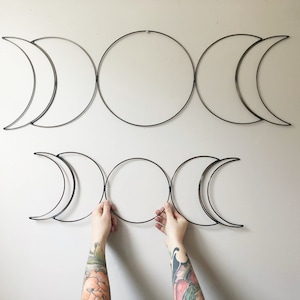 May include: Two sets of three metal moon phases in a crescent shape, hanging on a white wall. The larger set is above the smaller set. A person's hands are holding the smaller set of moon phases.