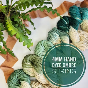 Peut inclure: Quatre pelotes de fil à tricoter ombré teintes à la main dans des tons de vert et crème. Le fil est disposé en cercle avec une étiquette qui indique "4MM HAND DYED OMBRE STRING".