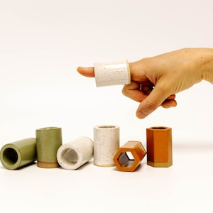 May include: A set of six ceramic shot glasses in various colours and shapes, including a hexagonal glass. The glasses are arranged in a row on a white surface.