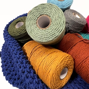 May include: A blue crocheted bag filled with various coloured yarn spools. The yarn shades include teal, olive green, grey, orange, and mustard yellow. The yarn is tightly wound on cardboard spools, ideal for crafting.