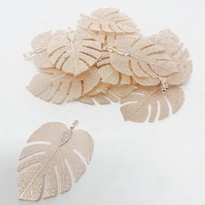 May include: A pile of gold-coloured metal monstera leaf charms. The leaves are delicate and have a detailed vein pattern. The charms have a loop at the top for attaching to jewellery or other crafts.