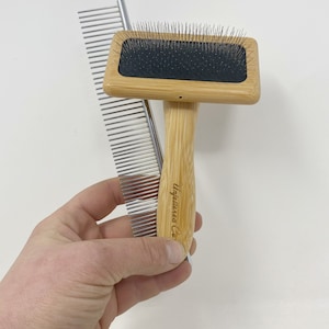 May include: A wooden pet grooming brush with a black bristle head and a silver comb. The brush has a wooden handle with the text "Unleashed Co." engraved on it.
