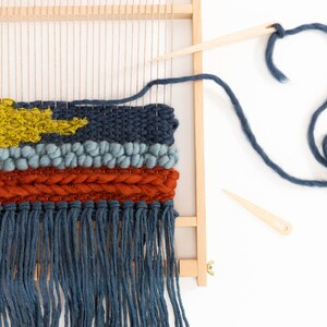 K&ouml;nnte beinhalten: Nahaufnahme einer gewebten Wandbeh&auml;nge mit einem Farbschema in Blau, Orange und Gelb. Die Beh&auml;nge befindet sich auf einem Holzwebstuhl mit blauem Garn und einer Holznadel.