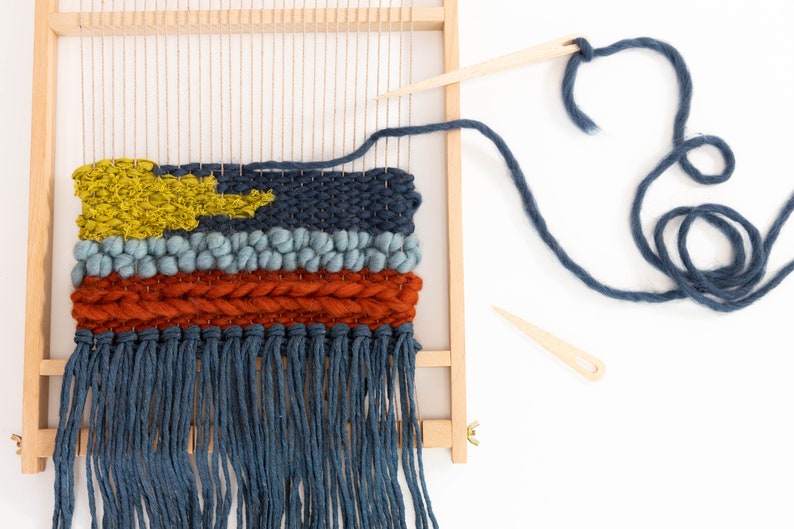 K&ouml;nnte beinhalten: Nahaufnahme einer gewebten Wandbeh&auml;nge mit einem Farbschema in Blau, Orange und Gelb. Die Beh&auml;nge befindet sich auf einem Holzwebstuhl mit blauem Garn und einer Holznadel.
