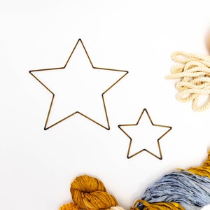 May include: Two gold metal star-shaped hoops, one larger than the other, on a white background. A pile of blue, yellow, and white yarn is in the corner of the image.