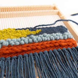 K&ouml;nnte beinhalten: Eine Nahaufnahme einer gewebten Wandbeh&auml;nge mit einem Holzrahmen. Die Beh&auml;nge weist eine Vielzahl von Texturen und Farben auf, darunter Blau, Gr&uuml;n, Orange und Gelb. Die Beh&auml;nge hat einen Fransen aus blauem Garn.