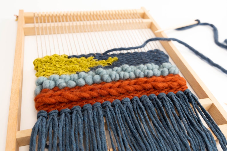 K&ouml;nnte beinhalten: Eine Nahaufnahme einer gewebten Wandbeh&auml;nge mit einem Holzrahmen. Die Beh&auml;nge weist eine Vielzahl von Texturen und Farben auf, darunter Blau, Gr&uuml;n, Orange und Gelb. Die Beh&auml;nge hat einen Fransen aus blauem Garn.