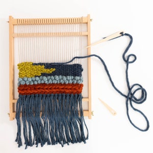 K&ouml;nnte beinhalten: Ein Holzwebstuhl mit einem gewebten Wandteppich mit einem gelben, blauen und roten Design. Der Wandteppich hat unten einen Fransen und ist teilweise fertig. Ein blaues Garn ist am Webstuhl befestigt und eine Holznadel liegt auf dem Tisch.