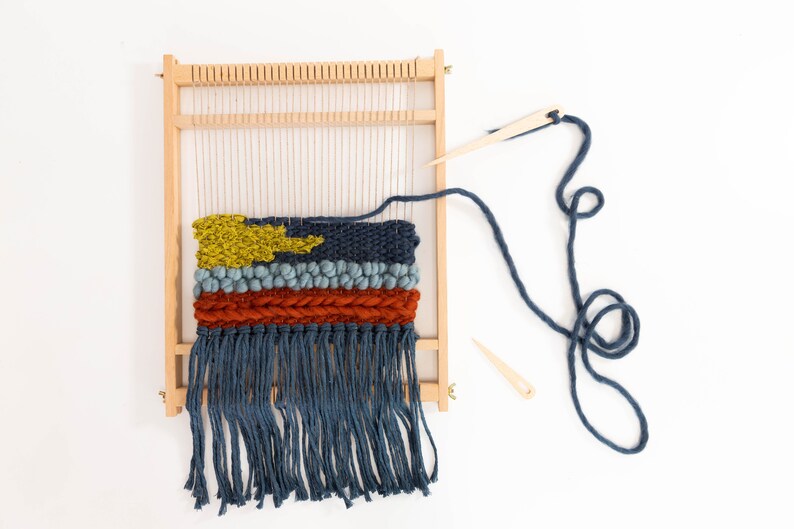 K&ouml;nnte beinhalten: Ein Holzwebstuhl mit einem gewebten Wandteppich mit einem gelben, blauen und roten Design. Der Wandteppich hat unten einen Fransen und ist teilweise fertig. Ein blaues Garn ist am Webstuhl befestigt und eine Holznadel liegt auf dem Tisch.