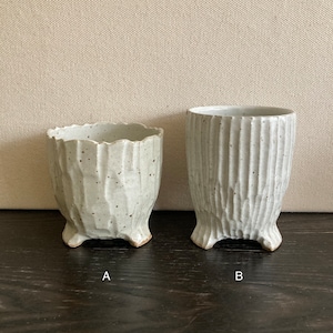 May include: Two white ceramic planters with a textured, rough surface and three legs each. The planter on the left is smaller than the planter on the right.