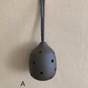 May include: A dark brown, rounded ceramic vessel with a textured surface and multiple circular cutouts, suspended by a thick grey rope loop. The letter A is printed in black to the lower left of the object, which hangs from a small metal hook.