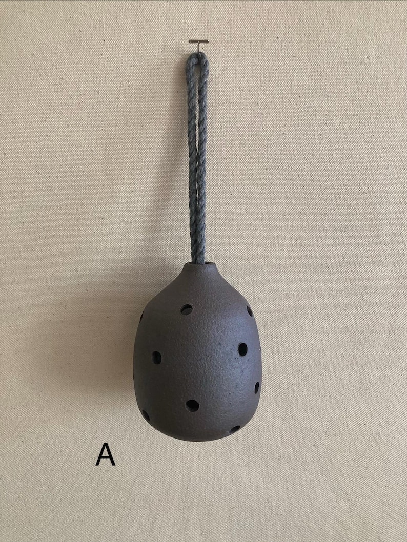 May include: A dark brown, rounded ceramic vessel with a textured surface and multiple circular cutouts, suspended by a thick grey rope loop. The letter A is printed in black to the lower left of the object, which hangs from a small metal hook.