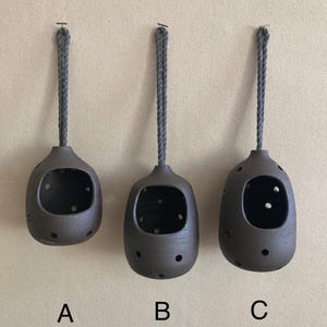 May include: Three dark brown ceramic hanging vessels labeled A, B, and C. Each vessel has a rounded shape, a large front opening, and multiple small circular perforations throughout the body. A grey braided rope is attached to the top of each.