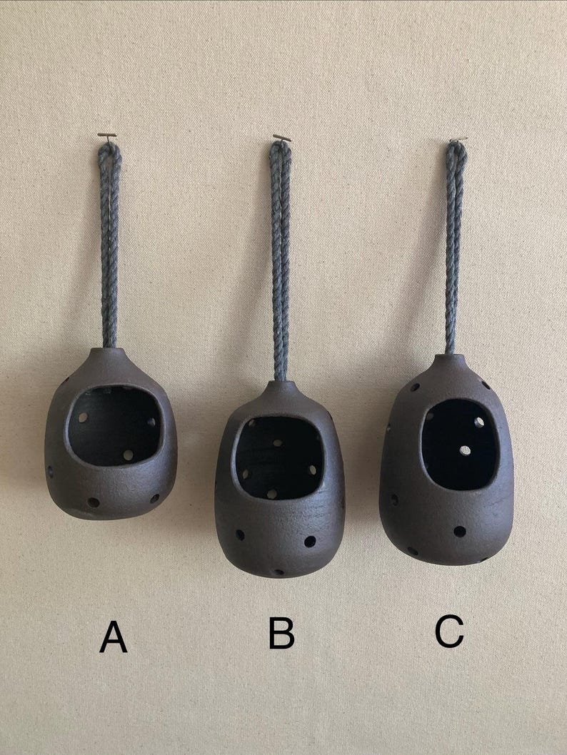 May include: Three dark brown ceramic hanging vessels labelled A, B, and C. Each vessel has a rounded shape, a large front opening, and multiple small circular perforations throughout the body. A grey braided rope is attached to the top of each.