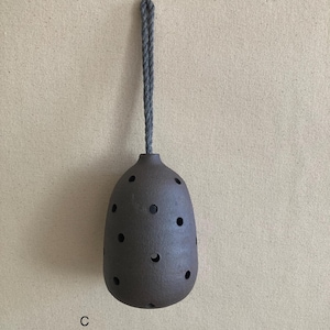 May include: A dark brown, egg-shaped ceramic vessel with a textured surface and multiple circular cutouts distributed across its body. A thick, grey, twisted rope cord is attached to the top for hanging. The letter C is printed in black.