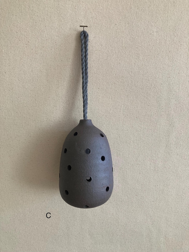 May include: A dark brown, egg-shaped ceramic vessel with a textured surface and multiple circular cutouts distributed across its body. A thick, grey, twisted rope cord is attached to the top for hanging. The letter C is printed in black.