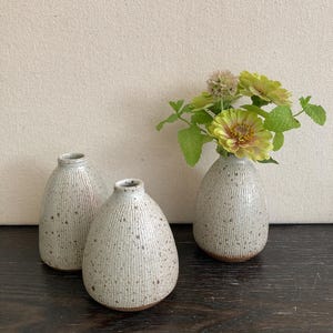 May include: Three small, speckled ceramic vases with a textured, vertical-line design. One vase holds a small bouquet of green and yellow flowers. The vases are on a dark wooden surface against a neutral background.