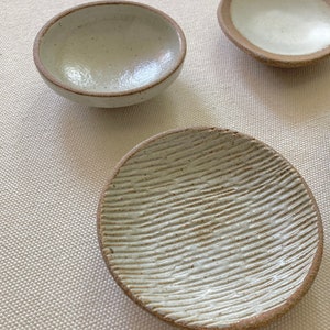 May include: Three white ceramic dishes with different textures. The dishes are round and shallow, with a slightly raised rim. The first dish has a smooth surface, the second dish has a textured surface, and the third dish has a smooth surface with a brown rim.