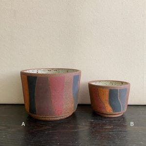 May include: Two ceramic pots with a rustic design. The larger pot features vertical stripes in black, brown, and red. The smaller pot has orange, black, and brown stripes. Both pots have a speckled interior and a dark brown exterior.