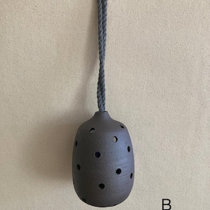 May include: A dark brown, rounded ceramic vessel featuring multiple circular cutouts across its surface. The object is suspended by a thick, grey twisted rope cord attached to the top opening. The letter B is printed in black in the lower right.