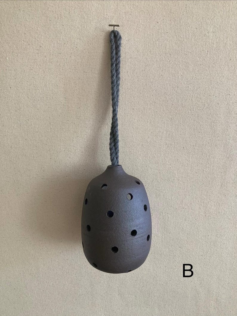 May include: A dark brown, rounded ceramic vessel featuring multiple circular cutouts across its surface. The object is suspended by a thick, grey twisted rope cord attached to the top opening. The letter B is printed in black in the lower right.
