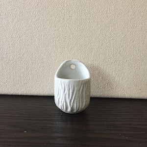 May include: White ceramic wall planter with a textured, wood-like design. The planter has a hole at the top for hanging.