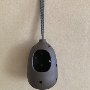 May include: A dark brown, rounded ceramic vessel with a large front opening and multiple smaller circular perforations distributed across its surface. The object is suspended by a thick, grey twisted rope loop attached to a small metal hook.