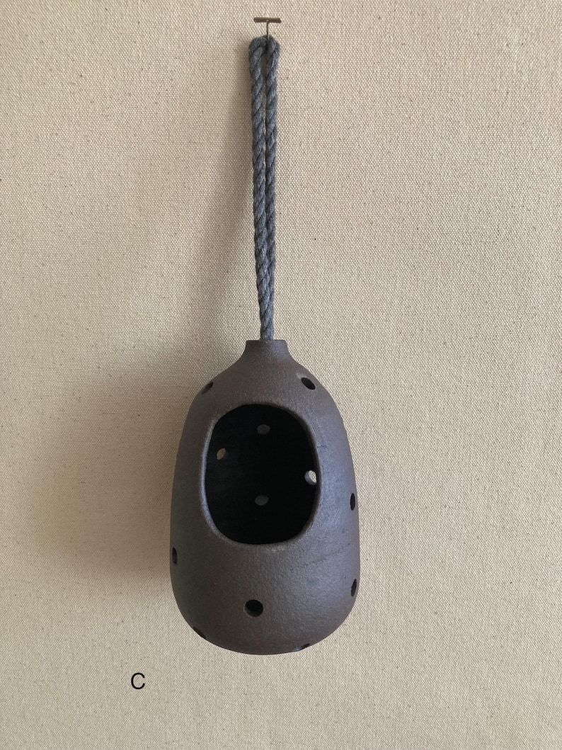 May include: A dark brown, rounded ceramic vessel with a large front opening and multiple smaller circular perforations distributed across its surface. The object is suspended by a thick, grey twisted rope loop attached to a small metal hook.