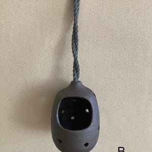 May include: A dark brown, rounded ceramic vessel with a large front opening and multiple small circular perforations along its sides. The vessel is suspended by a thick, twisted grey rope attached to a small metal hook at the top. The letter B is visible.