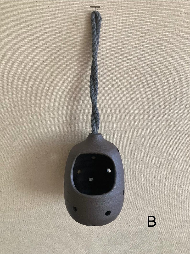May include: A dark brown, rounded ceramic vessel with a large front opening and multiple small circular perforations along its sides. The vessel is suspended by a thick, twisted grey rope attached to a small metal hook at the top. The letter B is visible.