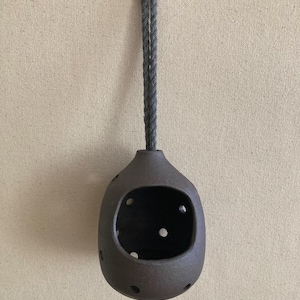 May include: A dark brown, rounded ceramic vessel with a large front opening and several smaller circular cutouts distributed across its surface. The vessel is suspended by a thick, grey twisted rope loop attached to a small metal hook.