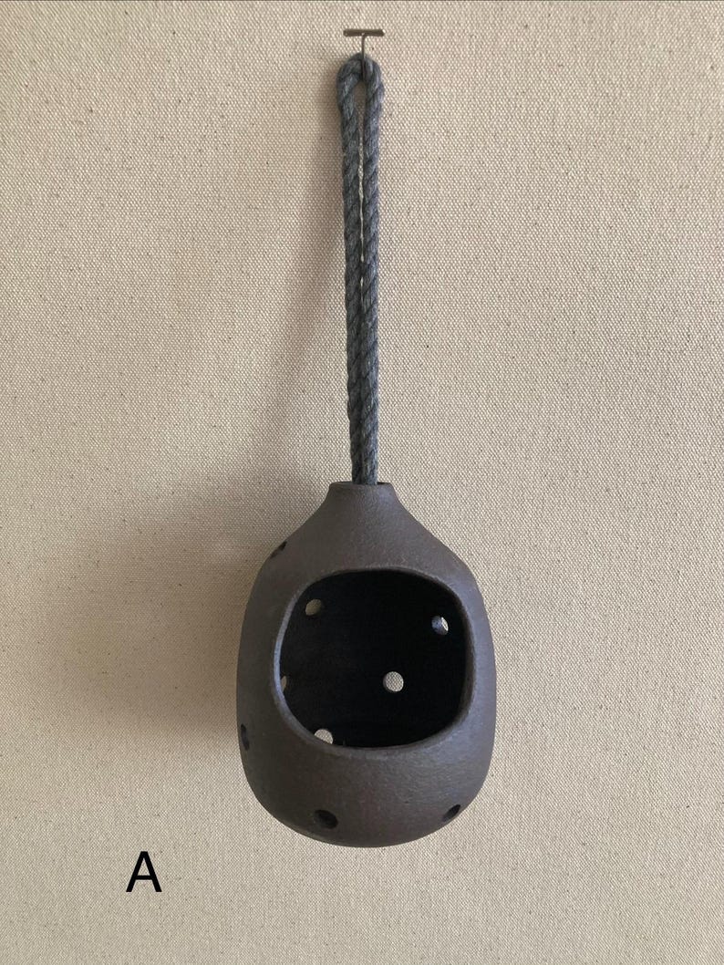 May include: A dark brown, rounded ceramic vessel with a large front opening and several smaller circular cutouts distributed across its surface. The vessel is suspended by a thick, grey twisted rope loop attached to a small metal hook.